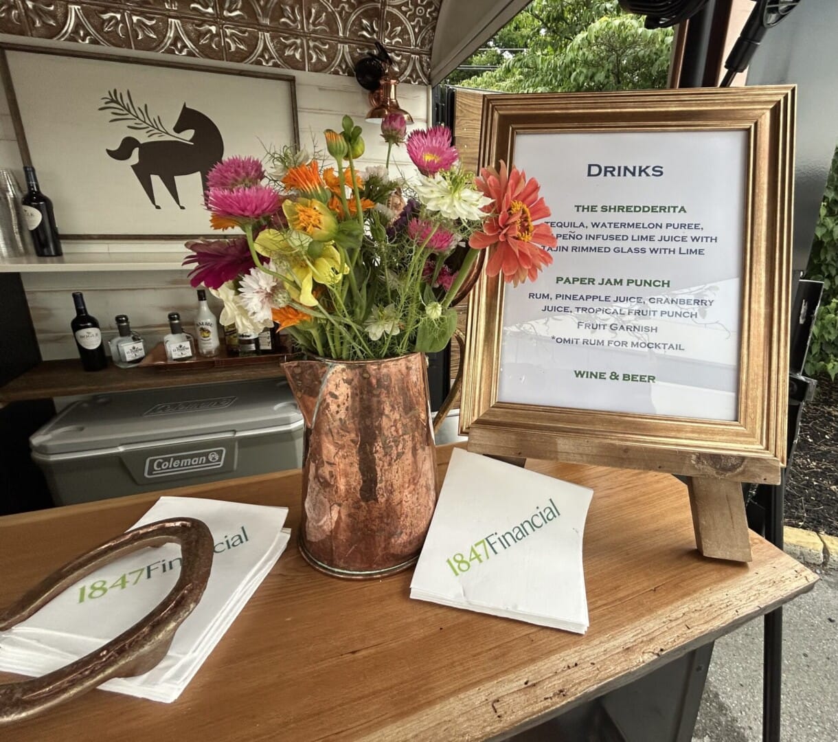 Copper Pony - A bar setup with a copper vase of colorful flowers, a framed drinks menu, and napkins labeled "1847 Financial" on a wooden counter. Bottles are visible in the background, creating a festive outdoor scene.