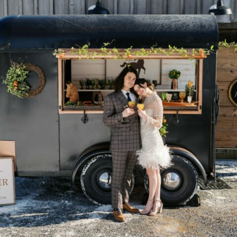 A couple stands in front of a stylish mobile bar trailer decorated with flowers and greenery. The man wears a plaid suit, and the woman wears a white dress. There's a "Just Married" sign on the bar, with a "Copper Pony" sign on the ground.