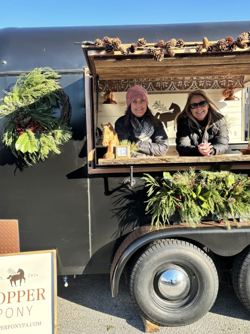 Copper Pony - Two women in winter clothing smile from the open window of a dark food truck decorated with greenery, pinecones, and festive wreaths. A sign reading "COPPER PONY" is visible next to the truck.