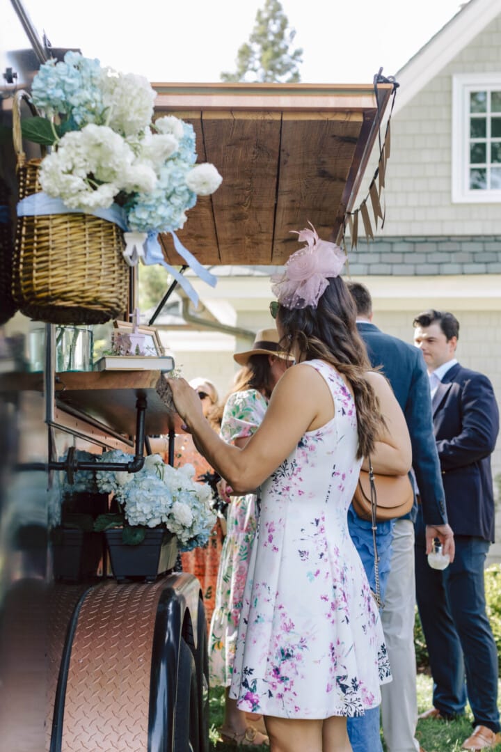 Copper Pony - A woman in a white floral dress stands at a flower-adorned outdoor vendor cart, with other people lined up behind her. The scene appears to be at a daytime garden event near a house.