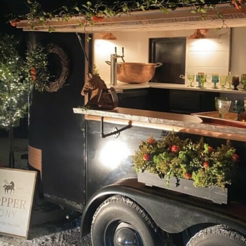 A converted horse trailer bar with plants and string lights adorns the exterior. Inside, a wooden counter, shelves with glasses, and a metal bowl are visible. The surrounding area features small trees with lights and a sign with text.