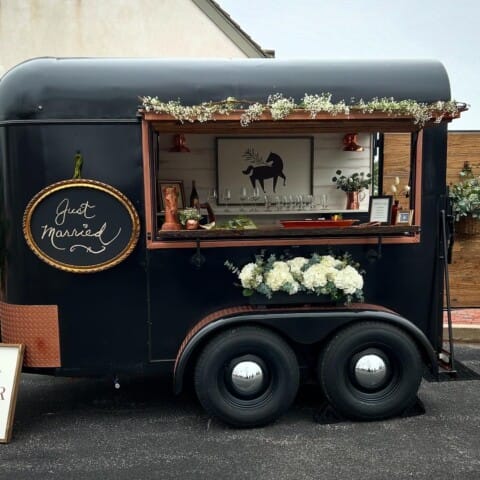 A black mobile bar trailer with floral decorations and a "Just Married" sign. It features a wooden counter with glasses and bottles. A logo reading "Copper Pony" is displayed below. The trailer is parked outdoors on a paved surface.