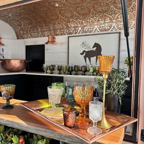 A countertop with various colored glasses on a copper tray. In the background, a metal ceiling pattern and a silhouette of a horse with foliage decorate the wall. A floral arrangement is visible in the foreground.