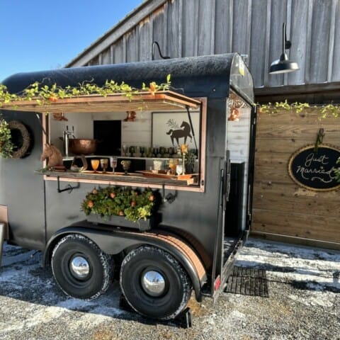 Mobile bar setup in a converted black trailer with a rustic vibe. The bar is decorated with greenery and copper elements. A wooden wall display reads "Just Married." It's parked on a snowy path near a rustic building.