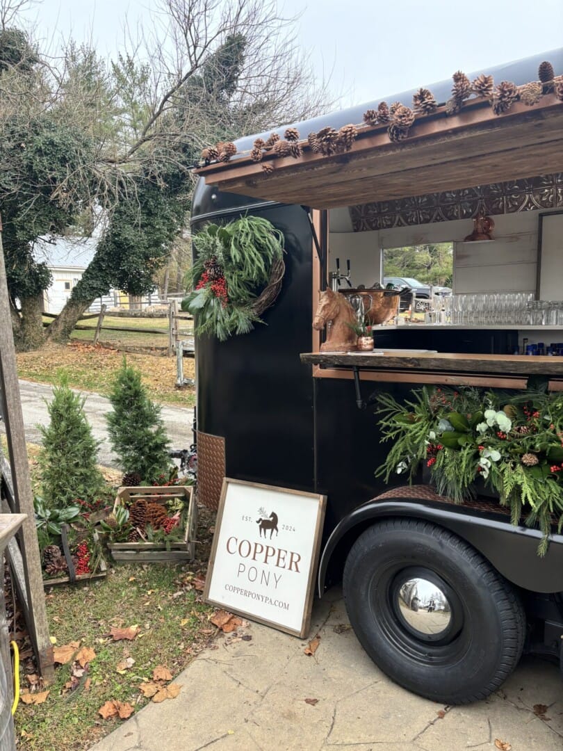 Copper Pony - A black truck decorated with pine cones, wreaths, and greenery serves as a mobile bar. A sign reading "Copper Pony" and small pine trees are placed beside the truck. Trees and grass are in the background.