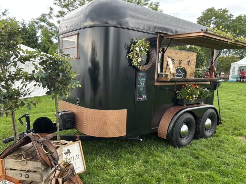 Copper Pony - A black food or drink trailer with a serving window is set up on grass. It’s decorated with plants and a wreath. Nearby are a saddle and a sign, and white tents are visible in the background.