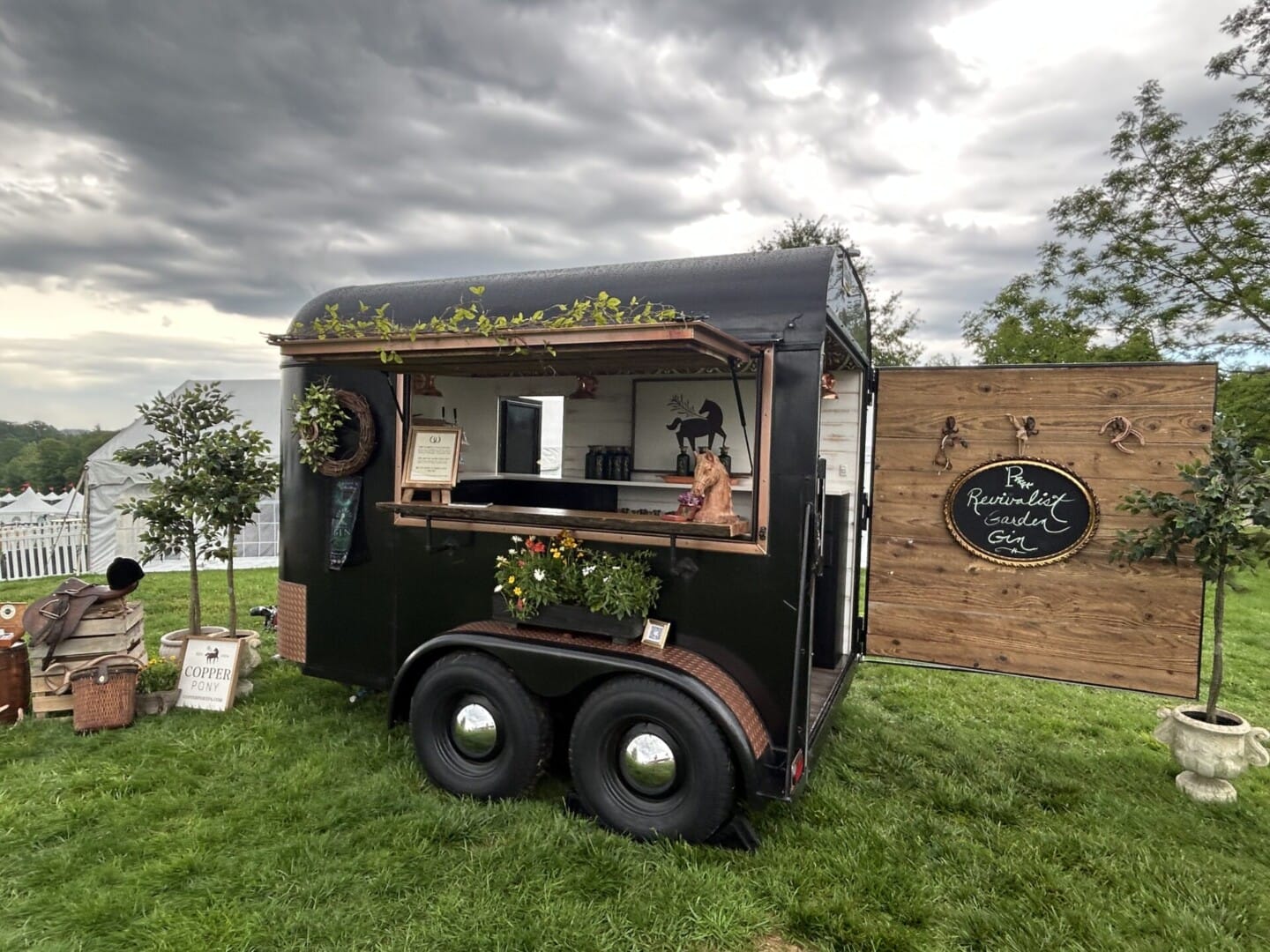 Copper Pony - A converted horse trailer serves as a rustic outdoor coffee and gin bar, set on grass with decorative greenery, a wooden sign, and picnic baskets nearby. Cloudy skies and a white tent are visible in the background.