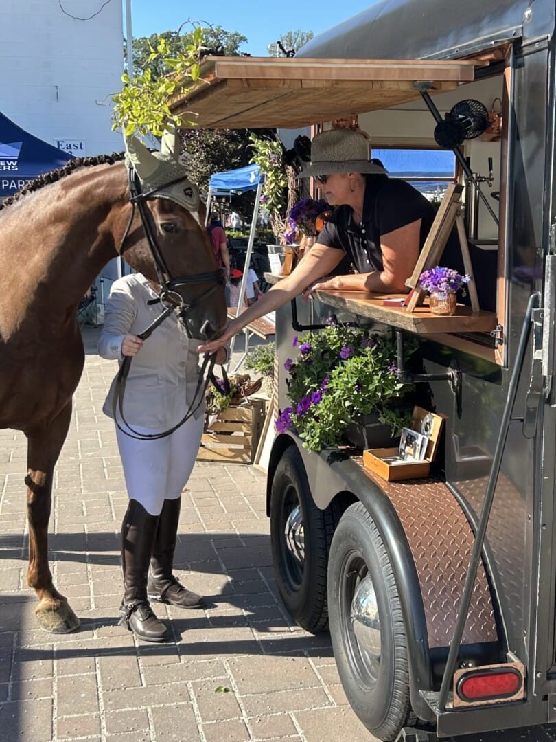 Copper Pony - A person in riding gear stands beside a brown horse at an outdoor market. The horse is reaching toward a vendor in a black hat serving from a trailer decorated with purple flowers and plants.