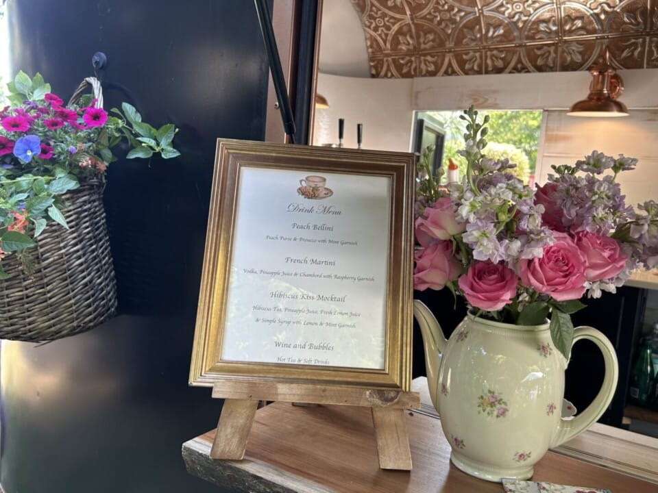 Copper Pony - A framed lunch menu sits on a small wooden easel next to a floral teapot vase with pink and purple flowers; a hanging basket of flowers and part of a kitchen are visible in the background.
