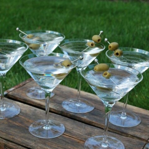 Copper Pony Vintage Mobile Bar Six martini glasses filled with clear liquid and garnished with green olives, each pierced with a toothpick. The glasses are arranged on a wooden surface against a backdrop of green grass.